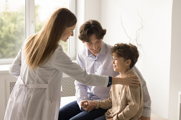 Fototapeta premium Young female pediatrician in white coat support little boy patient touch kid shoulder, give advice, provides medical assistance. Happy small child enjoy checkup meeting with dad. Check-up, healthcare