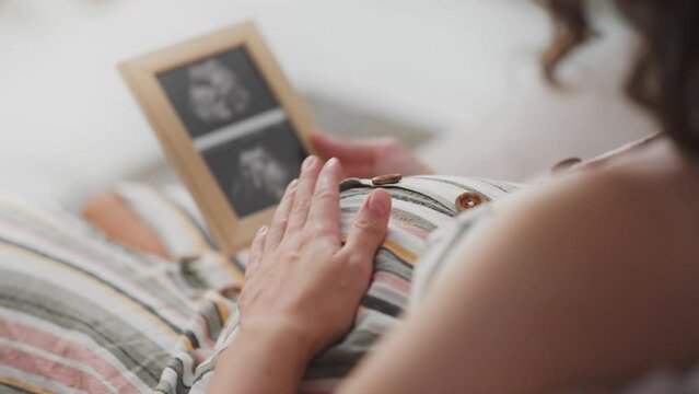 A pregnant girl looks at a photo of her unborn child's bridles in a room with bright light. A white woman holds a frame with a photo of a bridle in her hands, demonstrating concern for her unborn baby
