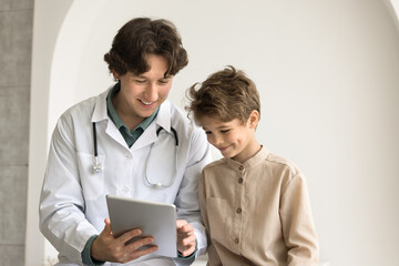 Pediatrician in white coat consulting to little 8s kid boy, showing good health test results, explain treatment plan use digital tablet during patient visit in private clinic. Health check-up, tech - obrazy, fototapety, plakaty