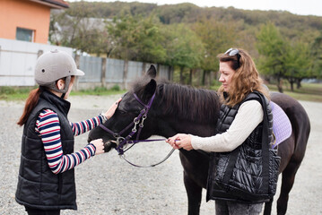A teenage girl after riding a horse in training and rehabilitation center, the girl needs hippotherapy after injury. A woman and her daughter are stroking a horse, try to make friends with it