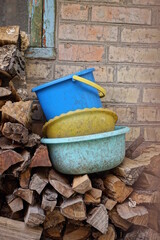 two old dirty yellow and blue bowls and one bucket stand on a gray pile of wooden logs near a brown brick wall on the street - obrazy, fototapety, plakaty