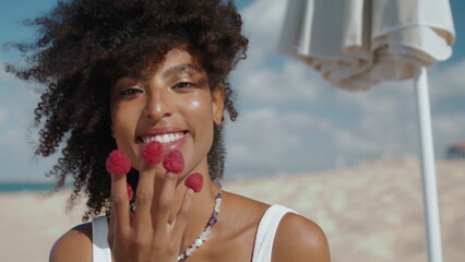 Beautiful girl eating raspberries of fingers closeup. Smiling woman rest beach