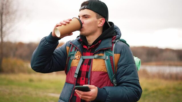 Young guy stands on outdoors nature landscape in forest with lake with hiking backpack on shoulders. Drink hot coffee for warming. Looking at screen of mobile phone, chatting in social networks.