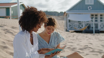 Two girls reading book on summer beach picnic. Happy lesbian couple talking