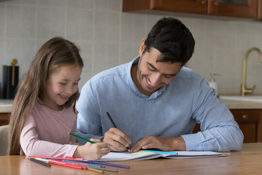 Happy little kid girl and handsome dad drawing colorful doodles in album together, using colored pencils, sitting at kitchen table, talking, laughing, having fun, training creative artistic skills