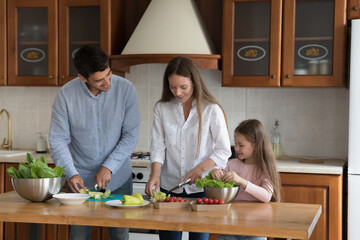 Cheerful parents and cute kid girl cooking salad together, preparing ingredients, slicing fresh vegetables at kitchen table, talking, laughing. Family household, healthy lifestyle concept