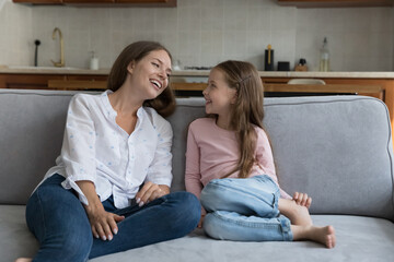 Happy sweet little kid girl and young pretty mom chatting on home sofa, laughing, resting on couch, enjoying family leisure time, funny talk, conversation. Motherhood, family, parenthood concept