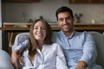 Obraz na płótnie Canvas Joyful happy attractive young couple video call shot portrait. Married man and woman resting on home sofa, hugging, looking at camera with toothy smiles, speaking, talking, laughing