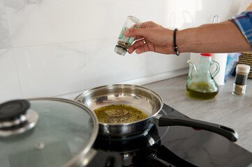Close-up of chef hand adding pepper into a frying pan with bay leaf and virgin olive oil on an electric stove