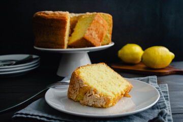 Lemon Pound Cake Slice on a Dessert Plate with a Fork: Plated slice of lemon pound cake with a whole cake on cake stand in the background
