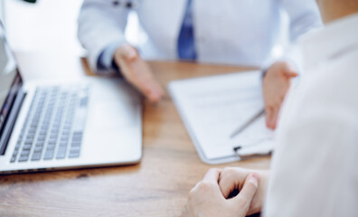 Doctor and patient sitting and discussing something at wooden table while using a laptop computer. Focus is on patient's hands. Medicine concept