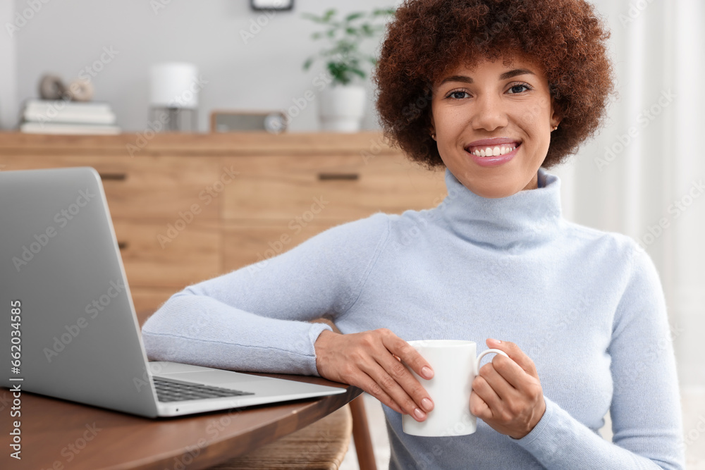 Wall mural Beautiful young woman using laptop and drinking coffee at wooden table in room