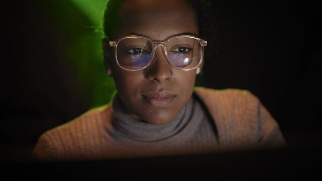 A young Latina business woman works late at computer. The laptop screen is reflected in the smiling girl glasses. Proud female worker at night. People concentrating on their projects on digital device