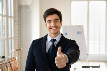Fototapeta premium Cheerful handsome young businessman in formal suit and tie posing in office boardroom with thumb up hand, making like sign, looking at camera with toothy smile, expressing satisfaction