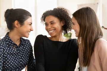 Multiethnic group of three cheerful employee girls standing close, talking, laughing, enjoying office friendship, communication, having fun. Happy young female colleagues, friends casual portrait