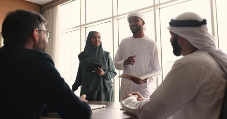 Positive Muslim teammates in traditional attire having friendly conversation with colleagues or company clients in conference room, smile, share ideas and creative solutions lead briefing in boardroom