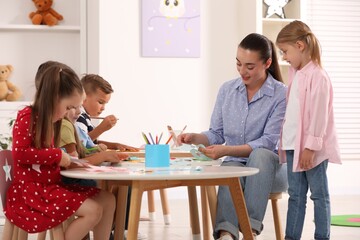 Nursery teacher and group of cute little children making toys from color paper at desks in kindergarten. Playtime activities