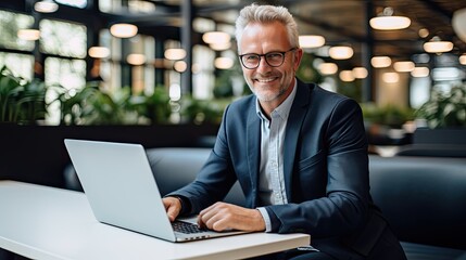 A professionally dressed man with white hair and glasses is seen seated in a contemporary office space. He is working on a sleek laptop placed on a white table, smiling at camera, 