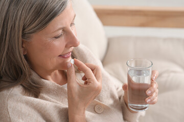 Senior woman with glass of water taking pill indoors, closeup