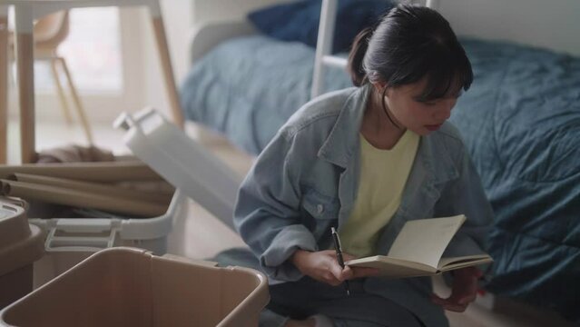 Happy young Asian Female A college student is creating the list of  carrying boxes in the notebook or smartphone before moving out of the dorm or to a new rented apartment room.