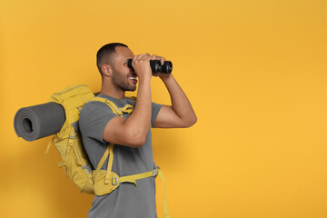 Happy tourist with backpack looking through binoculars on yellow background, space for text