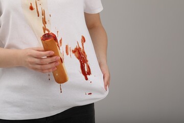Woman holding hotdog and showing stain from sauce on her shirt against light grey background, closeup. Space for text - obrazy, fototapety, plakaty