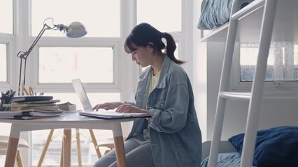 Asian girl student using laptop online learning class, study online preparing for school exams and making notes sitting at the desk in dormitory.
