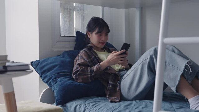 Happy young Asian Female A college student is using smartphone for chat on social network relaxing in comfortable bedroom at dormitory.