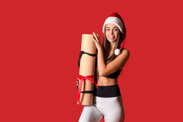 Sporty young woman in Santa hat with fitness mat as gift on red background