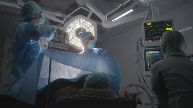 Male surgeons perform laparoscopy in modern well-equipped operating room. Surgical lamp shines on patient. Female medic checks condition of man on monitors. Medical staff at work in modern hospital.