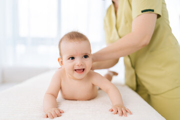 Portrait of happy smiling newborn baby lying on belly while unrecognizable female masseuse performing massage for small back and developing muscles. Mom hands doing back massage to newborn son.