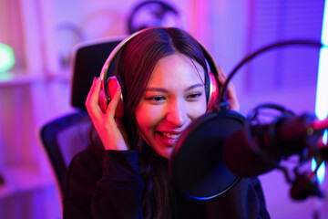 Asian podcaster girl saying tips and tricks in game online while using microphone and listening sound from wireless headphone gadget in technology channel and share on social media in radio station
