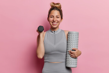 Horizontal shot of sportswoman with hair bun smiles broadly lifts dumbbell and holds foam roller smiles gladfully enjoys sport training dressed in grey activewear isolated over pink background