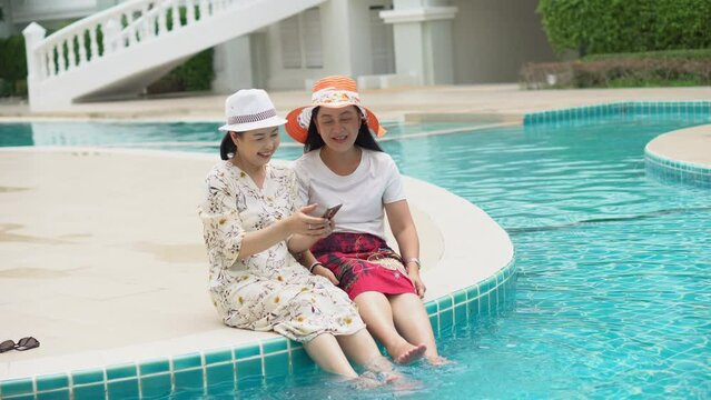 two senior asian woman with hat sitting by swimming pool take selfie with phone together outdoor. old lady take a photo . retired relaxation and vacation . friendship