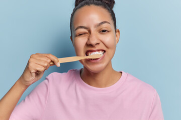 Horizontal shot of young Latin woman brushing teeth with toothbrush during morning time wears casual pink t shirt poses against blue background poses indoor. Dental care concept. Morning hygiene