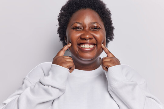Positive dark skinned chubbywoman points fingers at cheeks smiles toothily shows her happy emotions makes glad face dressed in casual sweatshirt isolated over white background has fake smile