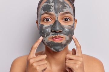 Headshot of surprised young Latin woman holds breath and blows cheeks applying facial clay mask for skin revitalization undergoes beauty treatments stands shirtless isolated over white background.