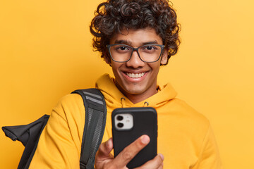 Photo of handsome curly haired Hindu man holds mobile phone in hand sends text messages wears transparent eyeglasses and casual hoodie carries backpack on shoulder isolated over yellow background