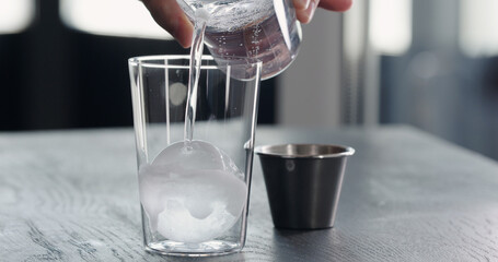 pour tonic water over clear ice ball in tumbler glass on black oak table