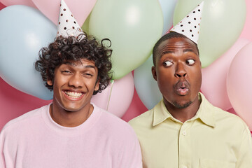 Studio photo of young cheerful smiling broadly Hindu and surprised African american men standing close to each other among colorful balloons celebrating birthday having party spending good time