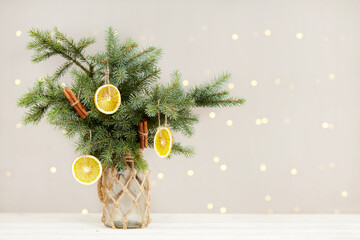 Winter bouquet of fir branches. Christmas tree in a vase with eco-friendly organic decor, dried orange and cinnamon sticks on the table against the background of bokeh lights. Zero waste concept. - obrazy, fototapety, plakaty