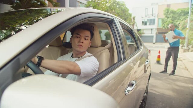 Asian man training reverse parking between cones while passing exam under supervision of driving instructor