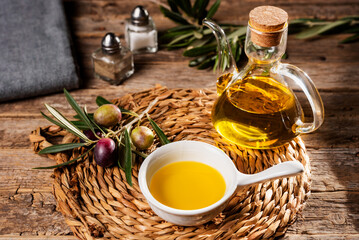 Small bowl with olive oil next to an oil can and some olives on a branch, rustic background.