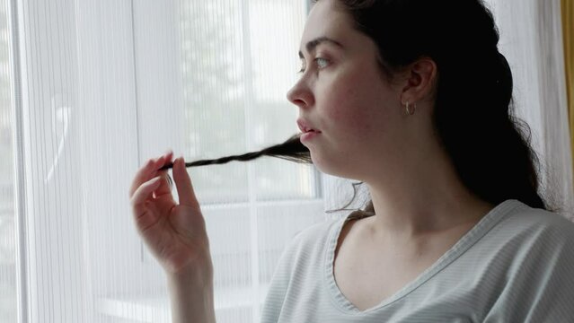 Anxious Caucasian woman curls lock of hair on her finger and lookind at window. Jib shot. Concept of psychological disorder ocd and mental problem