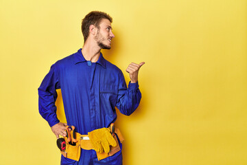 Man in blue jumpsuit with toolbelt on yellow points with thumb finger away, laughing and carefree.