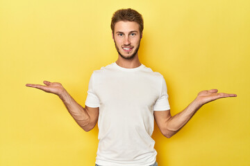 Young Caucasian man on a yellow studio background makes scale with arms, feels happy and confident.