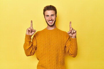 Young Caucasian man on a yellow studio background indicates with both fore fingers up showing a...