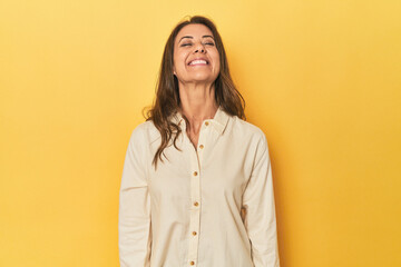 Middle-aged caucasian woman on yellow laughs and closes eyes, feels relaxed and happy. - Powered by Adobe