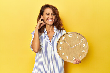 Middle aged woman holding a wall clock on a yellow backdrop covering ears with hands.