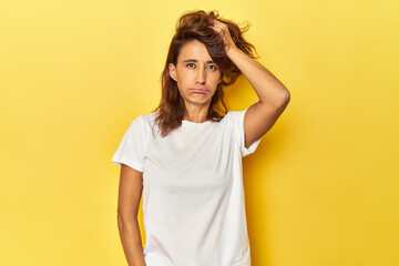 Middle-aged woman on a yellow backdrop tired and very sleepy keeping hand on head.